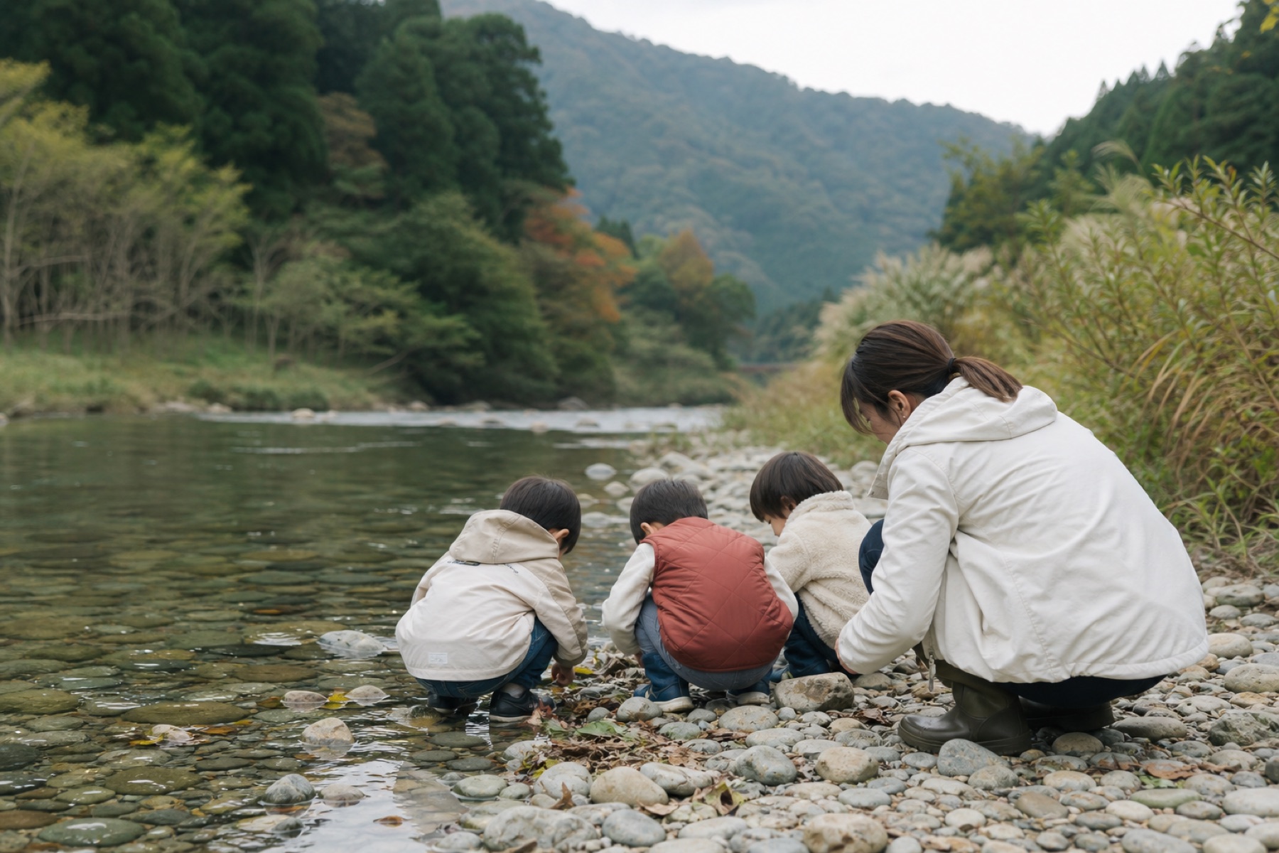山あいの川辺で、水際を一緒にのぞき込む子どもたちと保育者