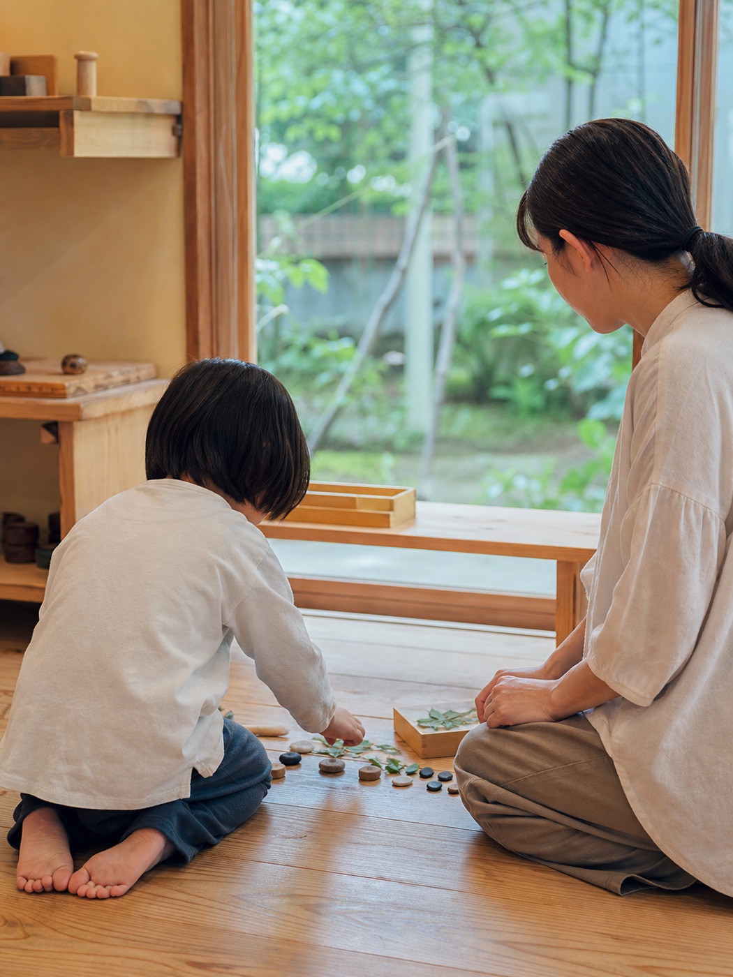 自然光の入る室内で、床に並べた石や葉を見る子どもと保育者
